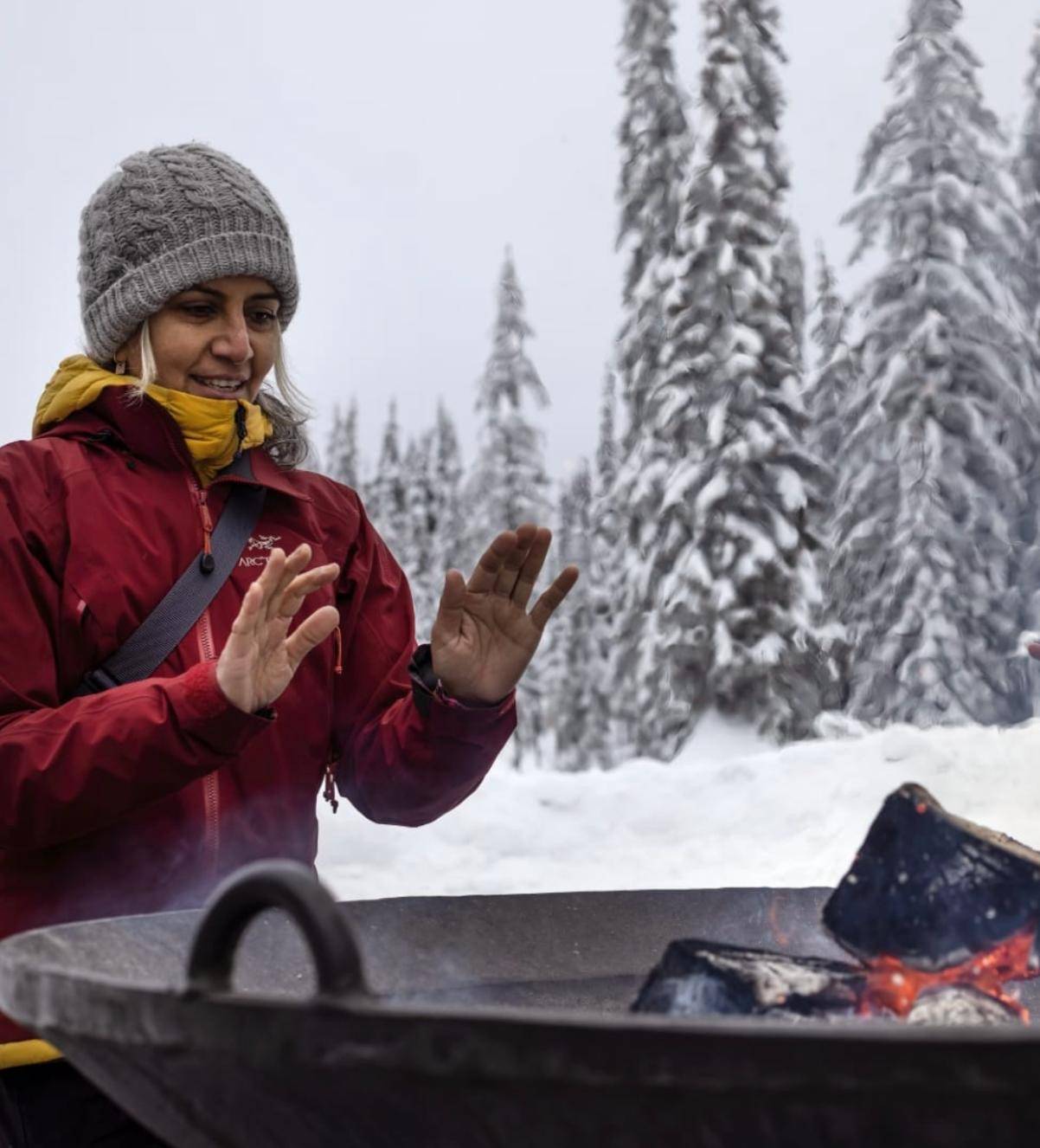 A person in a red jacket and gray beanie warms hands over an outdoor fire pit. Snow-covered trees in the background suggest a chilly, serene winter setting.