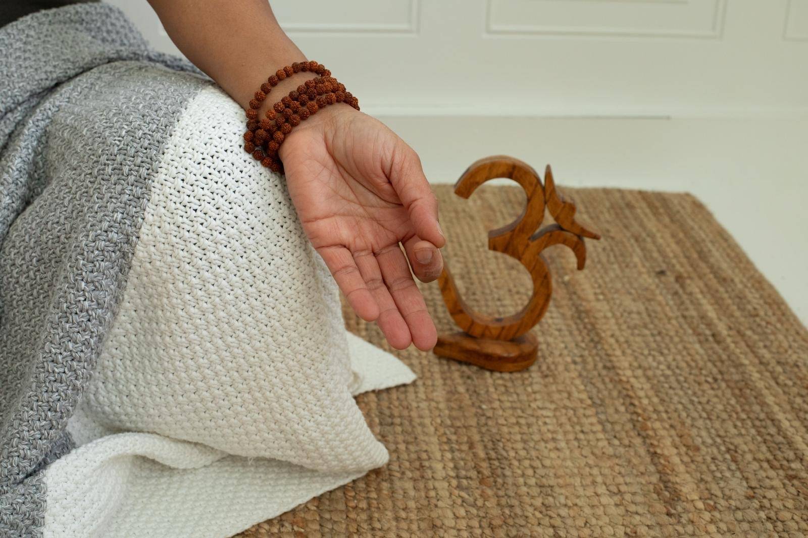 A person sits cross-legged on a woven mat, with their hand resting on their knee, fingers relaxed. A wooden Om symbol is in the background, evoking tranquility.