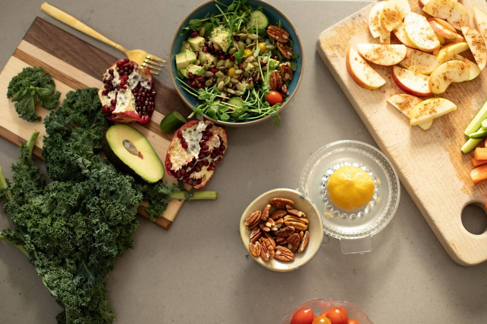 A colorful spread of fresh ingredients on a kitchen counter. Includes kale, avocado, pomegranate, salad bowl, pecans, lemon, apple slices, and carrots.