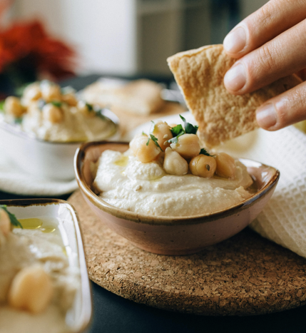 A hand holds a pita chip over a bowl of creamy hummus topped with chickpeas and parsley. Nearby, more bowls of hummus are arranged on a cork mat.