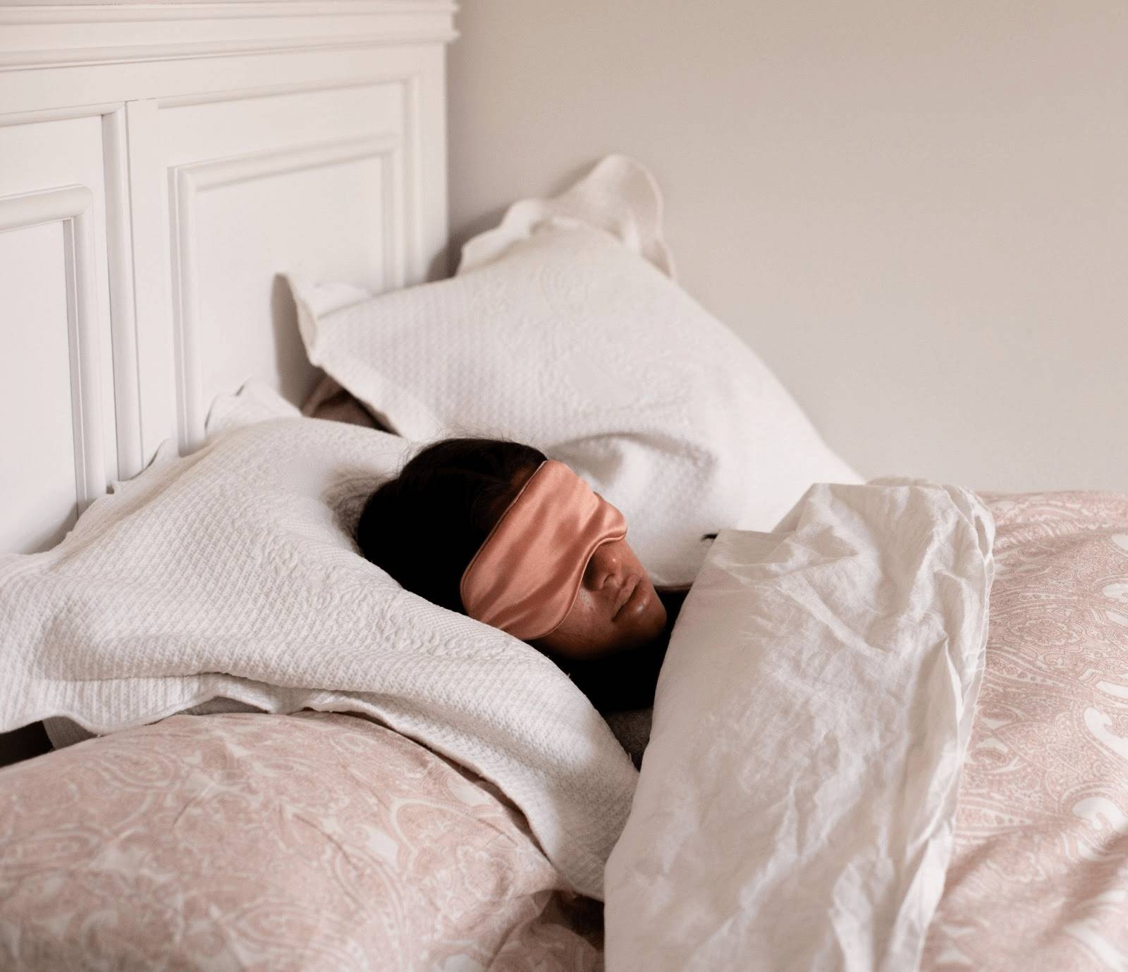 A person with a pink sleep mask lies peacefully in bed, wrapped in white sheets and pillows. The setting appears calm and cozy, suggesting restful sleep.