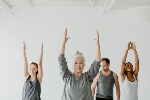 A diverse group of four people practice yoga indoors, standing with arms raised. They appear focused and peaceful, wearing gray and white athletic wear.