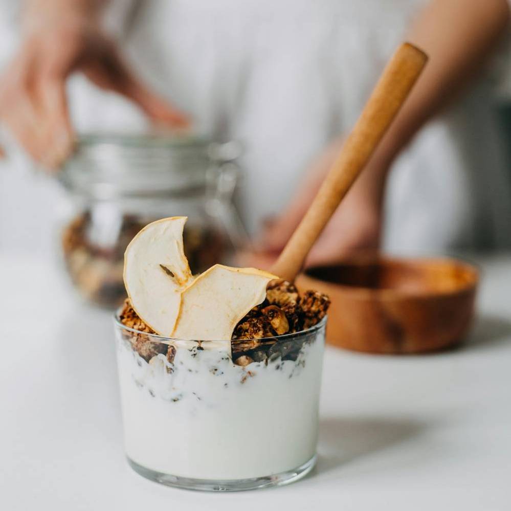 A glass of yogurt topped with granola and a thin apple slice, with a wooden spoon